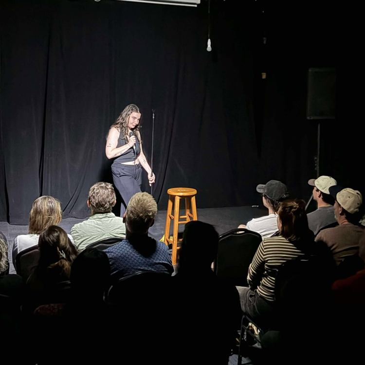 A comedian on stage beside the stool, in front of a full crowd at Club Voltaire